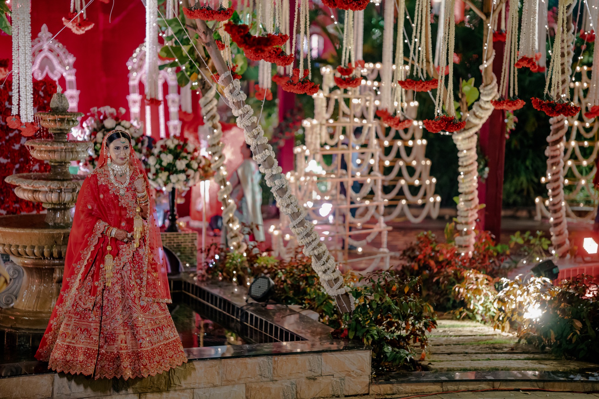 A bride in a red lehenga beside a fountain under marigold and jasmine garlands