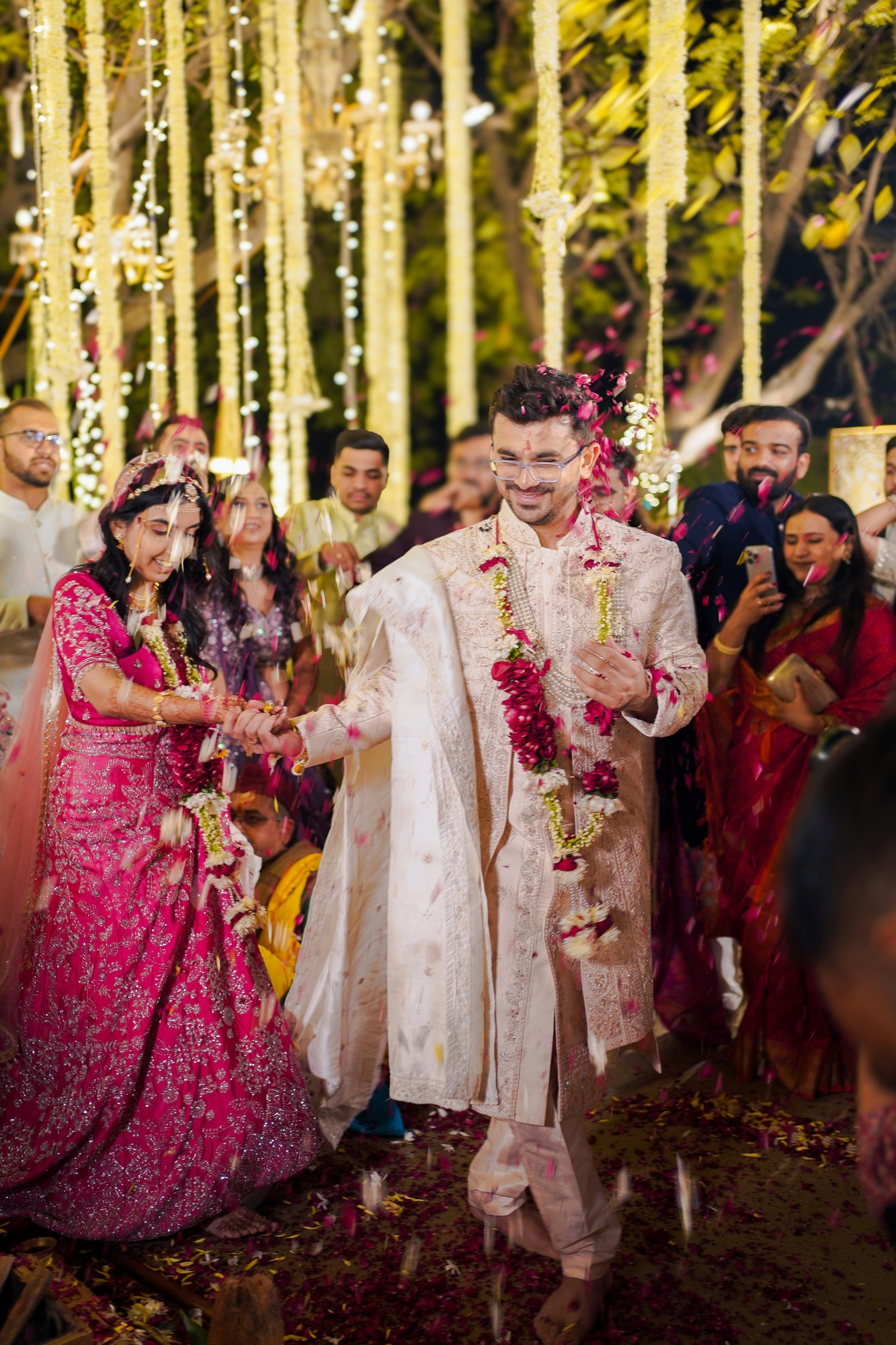 A bride in a pink lehenga and groom in an ivory sherwani walking hand in hand under a shower of rose petals, jasmine strings and fairy lights behind them