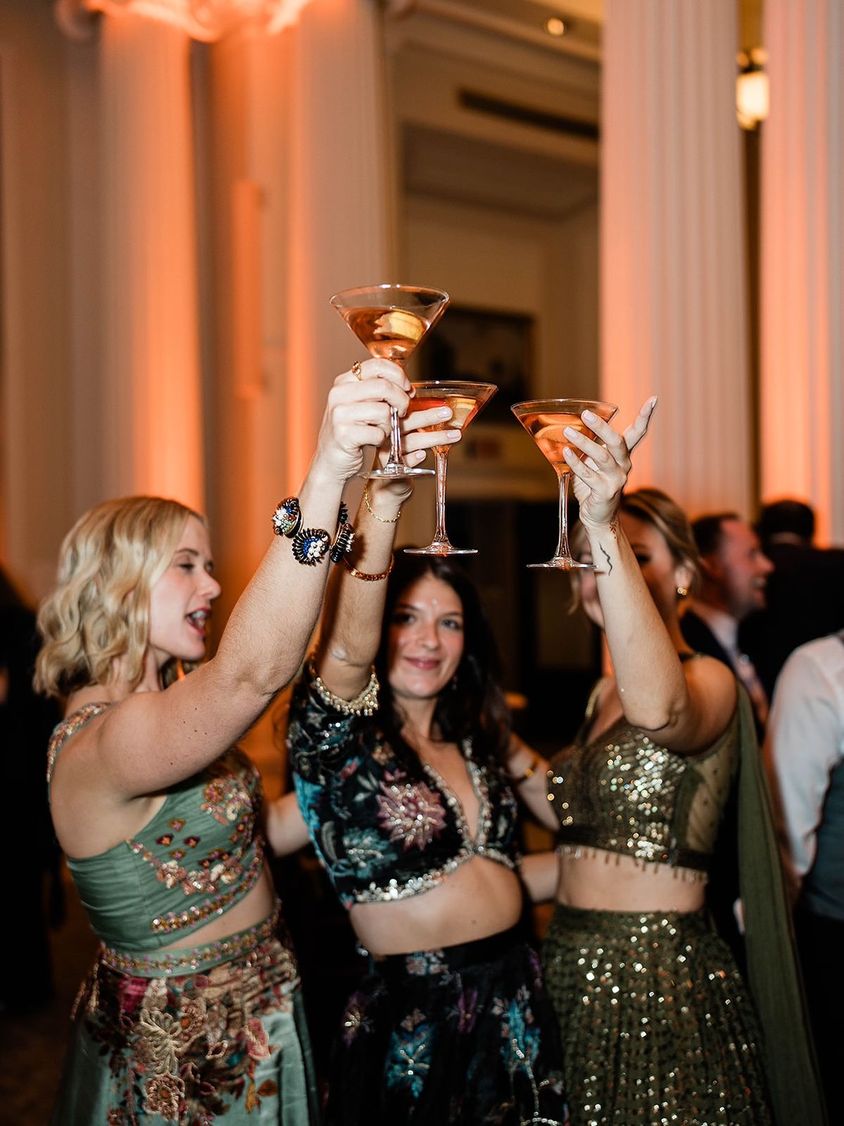 Three friends in embroidered lehengas raising martini glasses together at a cocktail party