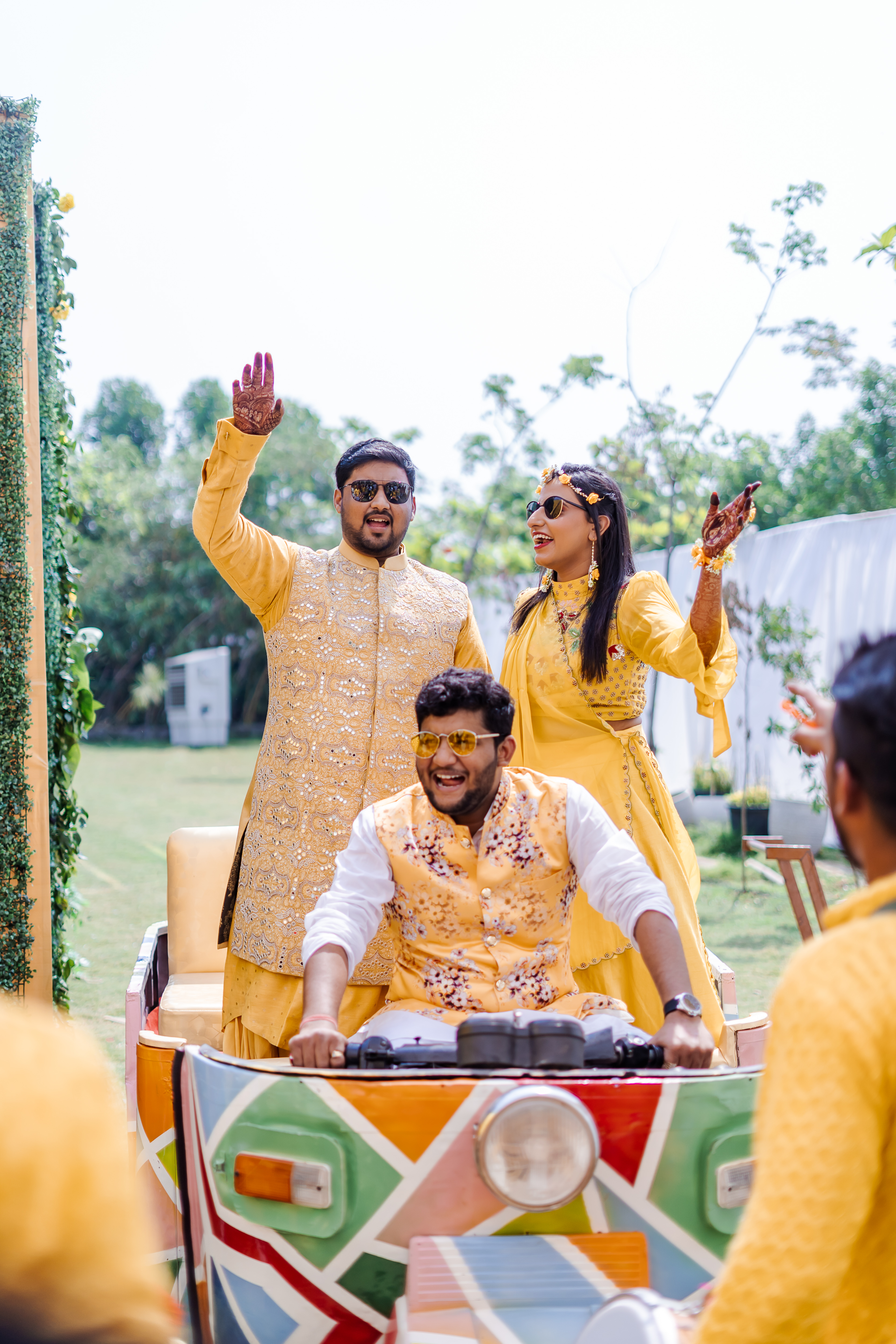 A couple in yellow on a hand-painted rickshaw at their haldi, hands raised mid-celebration