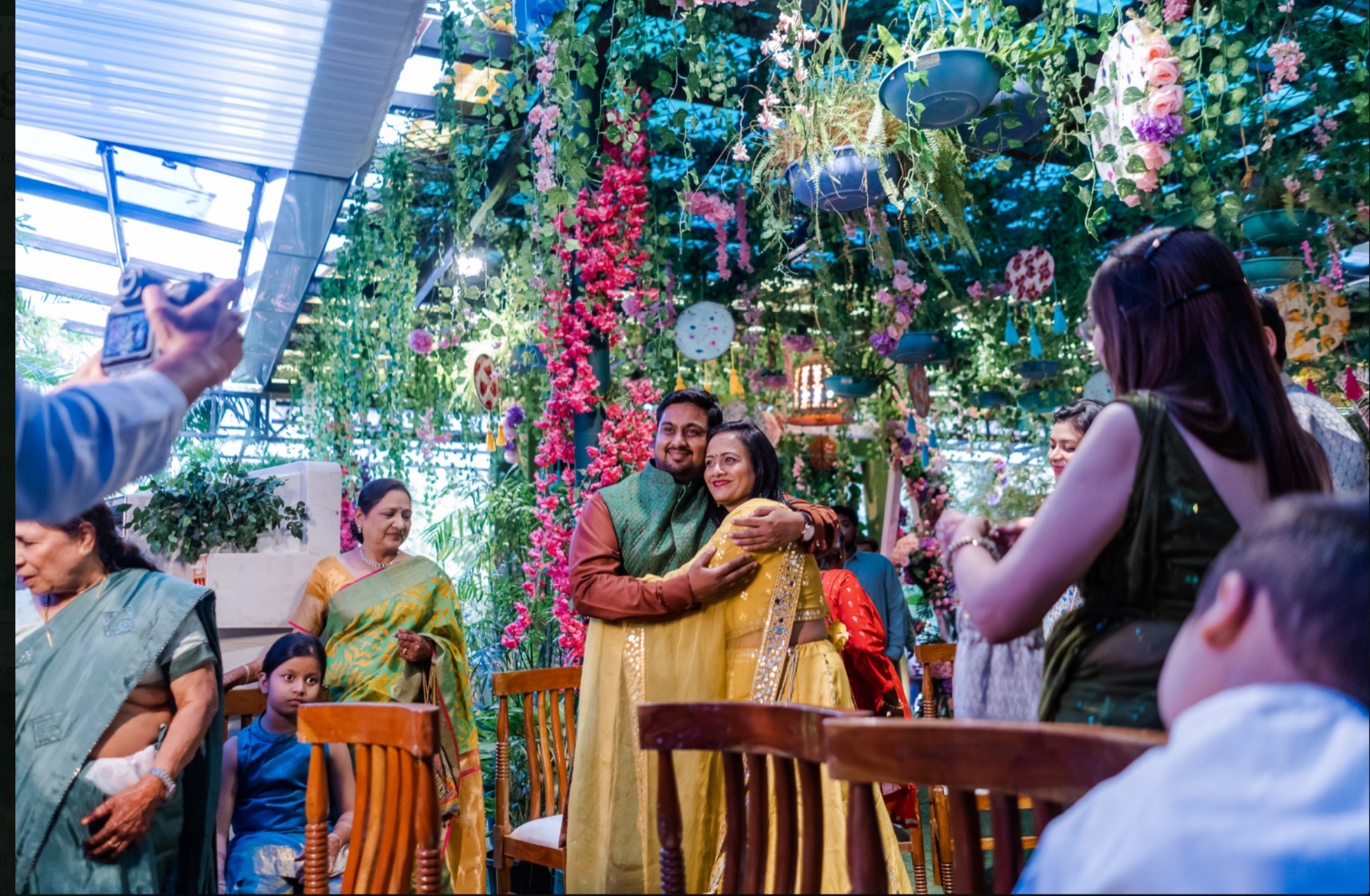 A groom embracing his mother under a flower-draped pergola as family looks on