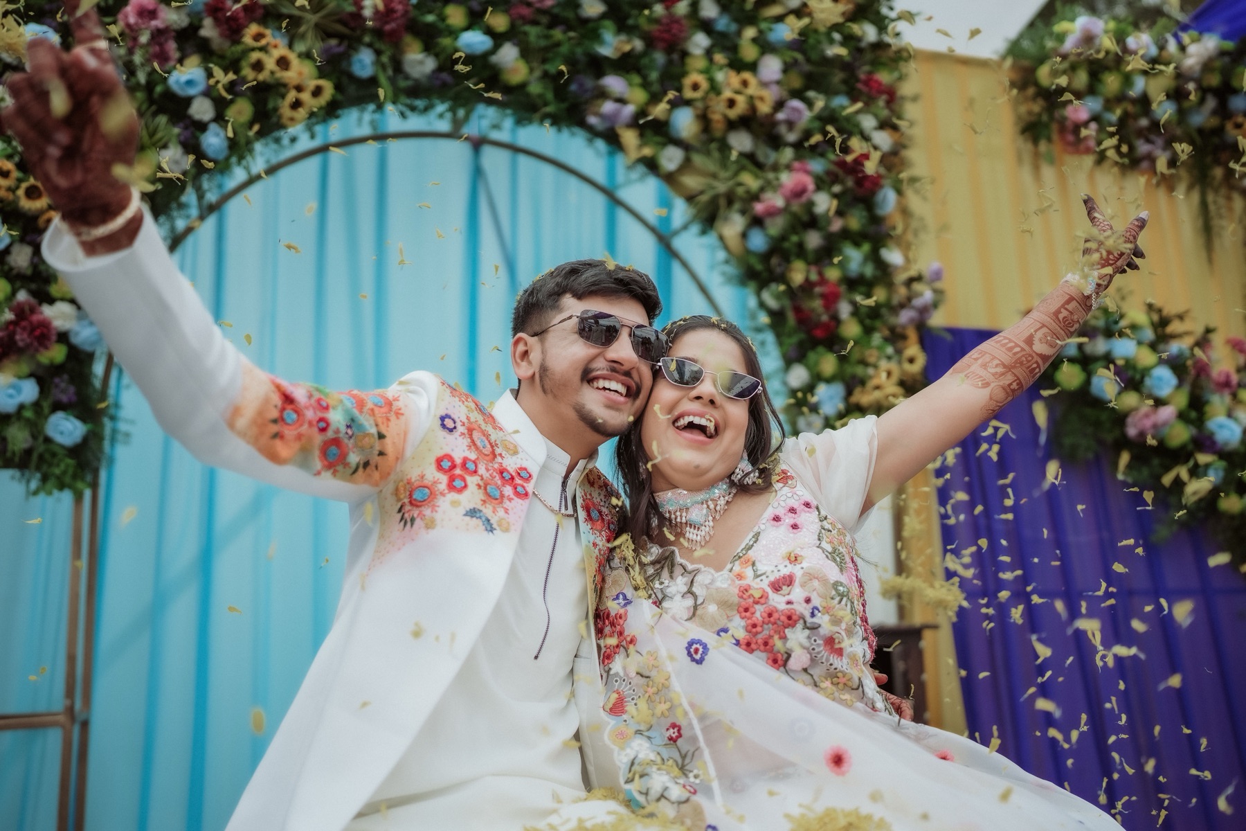 A laughing couple in sunglasses and embroidered white, arms flung wide as marigold petals shower down before a floral arch
