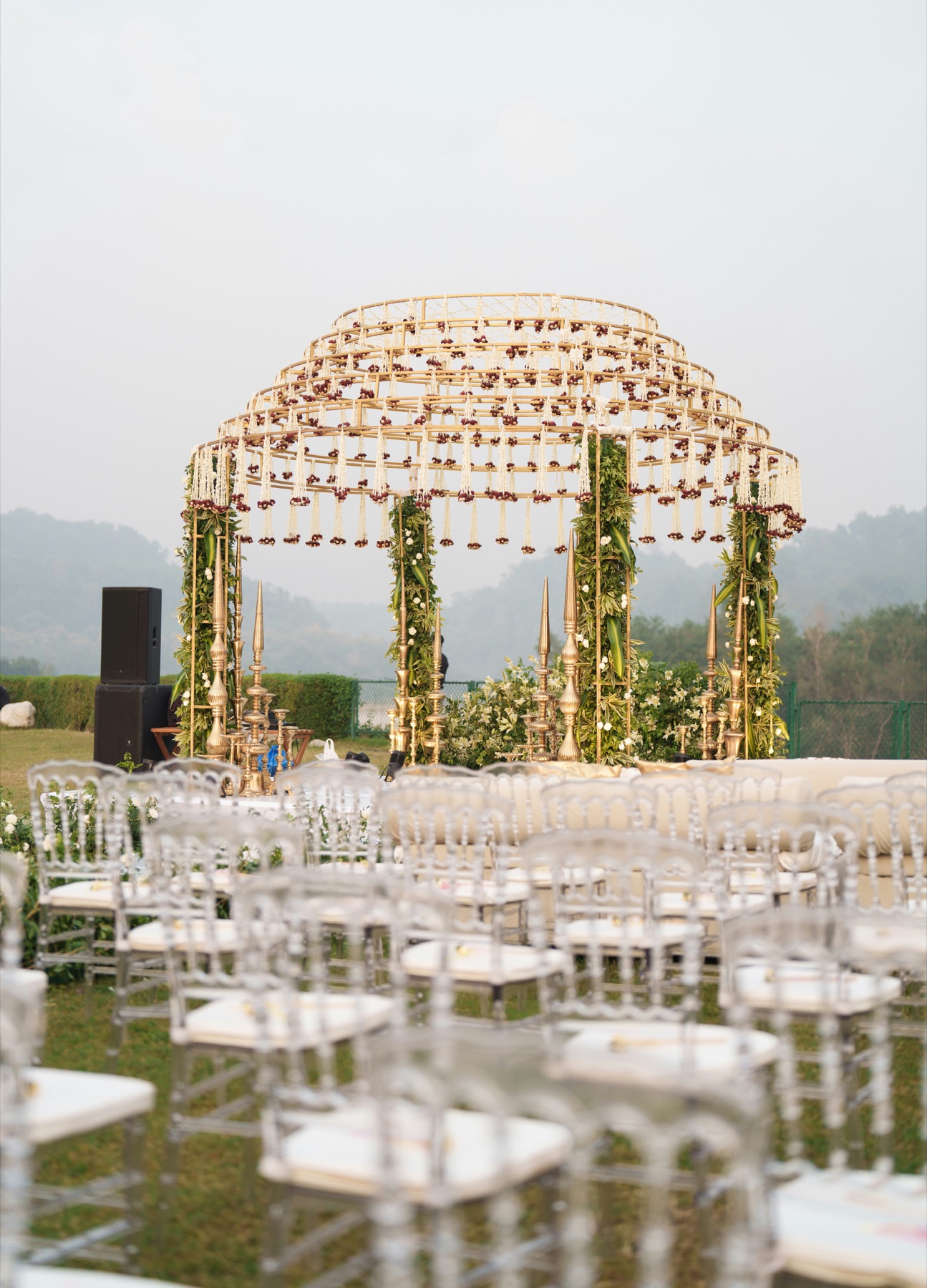 A gold ring mandap hung with jasmine and rose strands, set on a lawn before rows of clear chairs