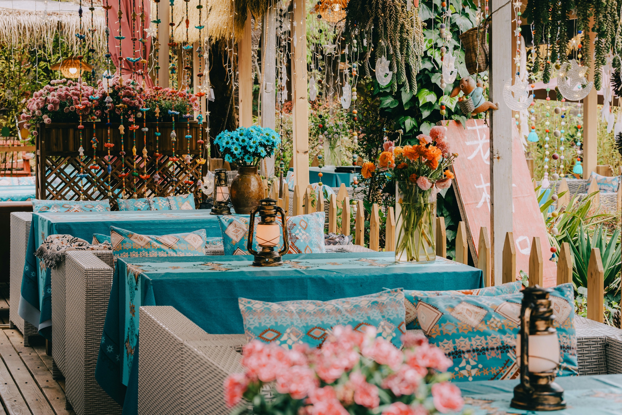 An outdoor lounge with teal tablecloths, patterned cushions, brass lanterns and fresh flowers under hanging beaded strings