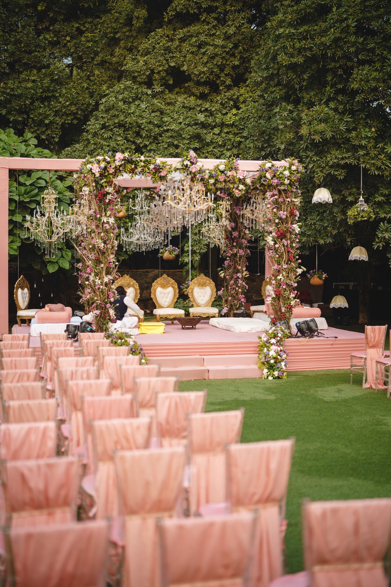 A flower-wrapped mandap with crystal chandeliers and gold chairs, facing rows of chairs draped in blush fabric on a lawn