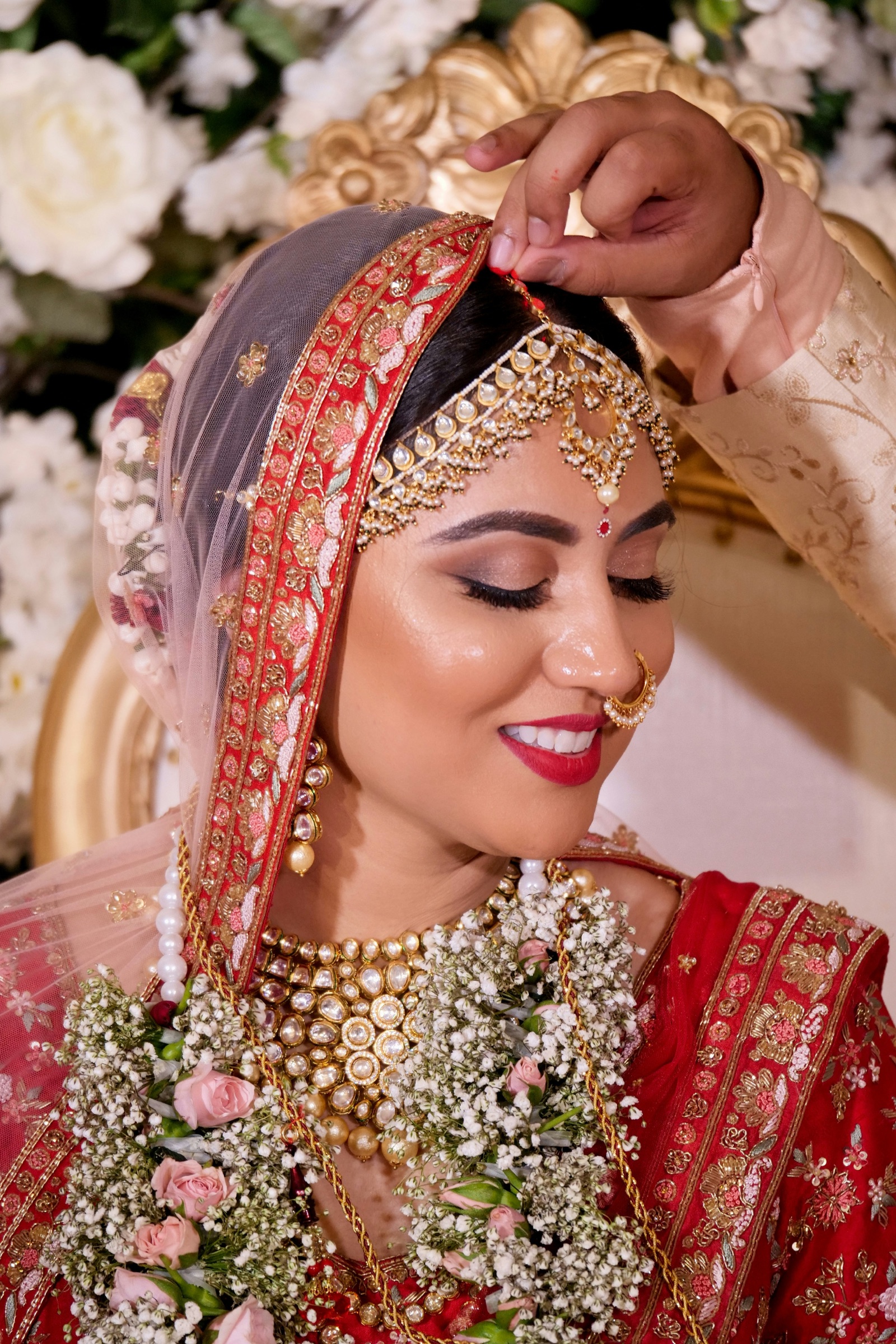 A bride in a red embroidered lehenga and kundan jewellery, smiling as her dupatta is adjusted
