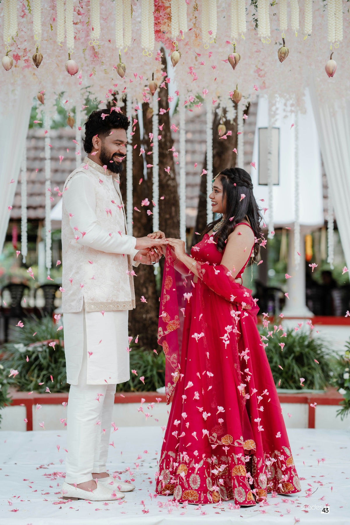 A couple exchanging rings under jasmine and lotus garlands as pink petals fall around them