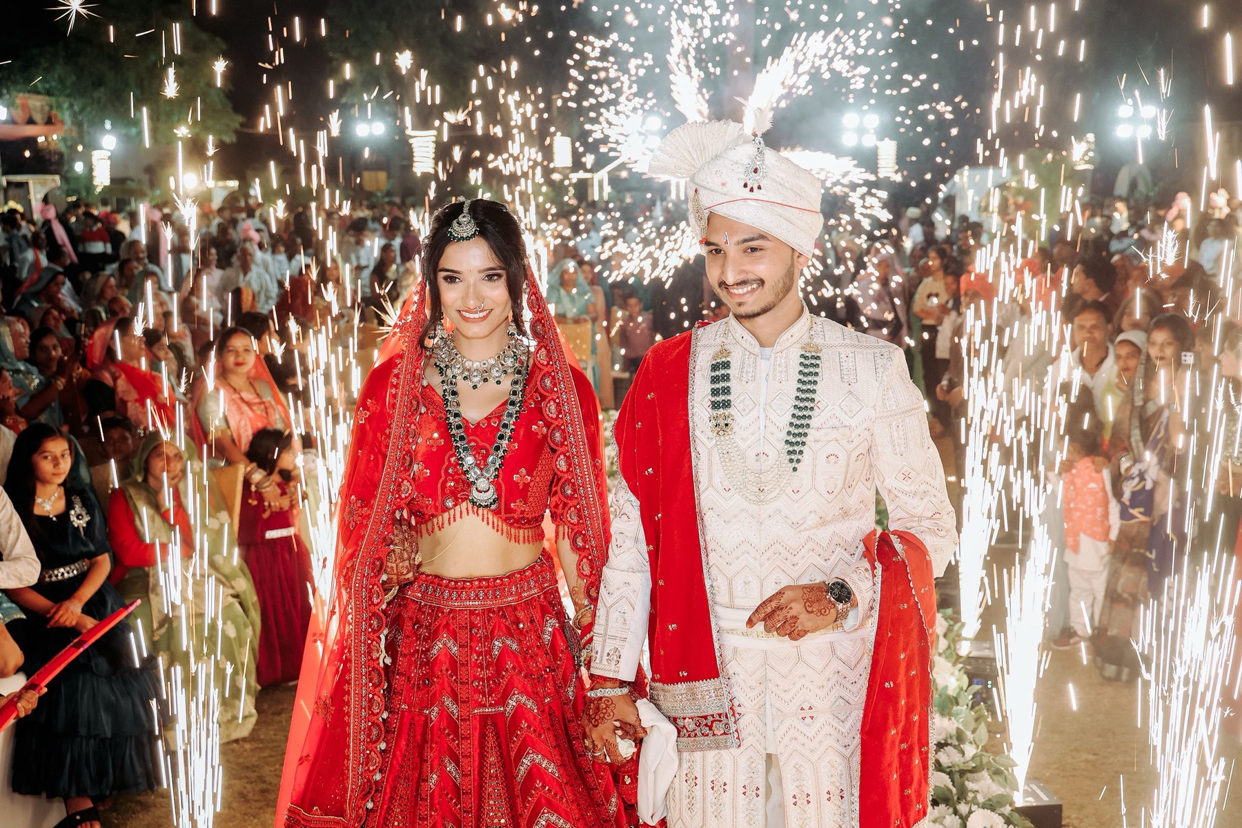 A smiling bride in a red lehenga and groom in an ivory sherwani walking hand in hand between fountains of sparks